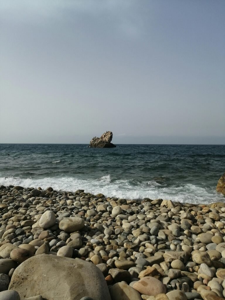 Explore the serene rocky beach with waves and distant rock formation in Béjaïa, Algeria.