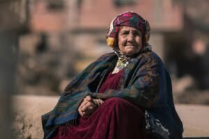 Elderly woman in Ghassira, Algeria, wearing traditional clothes and headscarf, sitting outdoors.